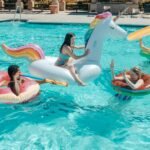 Group of adults enjoying a summer pool party with colorful inflatable floats and water guns.
