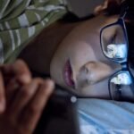A child lying in bed at night using a smartphone, with screen reflection on eyeglasses.